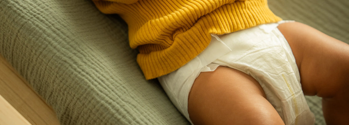 A close-up lifestyle shot of a baby wearing a white disposable diaper and a mustard yellow ribbed knit sweater, lying on a textured sage green blanket.